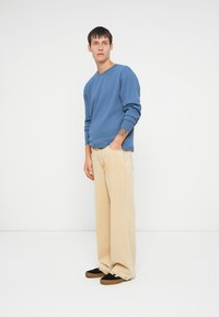 Sweatshirt azul de gola redonda, calças largas de um bege claro, ténis pretos com detalhes brancos, em pé contra um fundo branco liso.