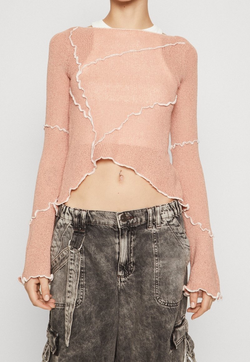 Pull en tricot rose court avec des bords volants, des manches longues et un ourlet blanc contrastant, associé à un pantalon cargo gris déchiré.