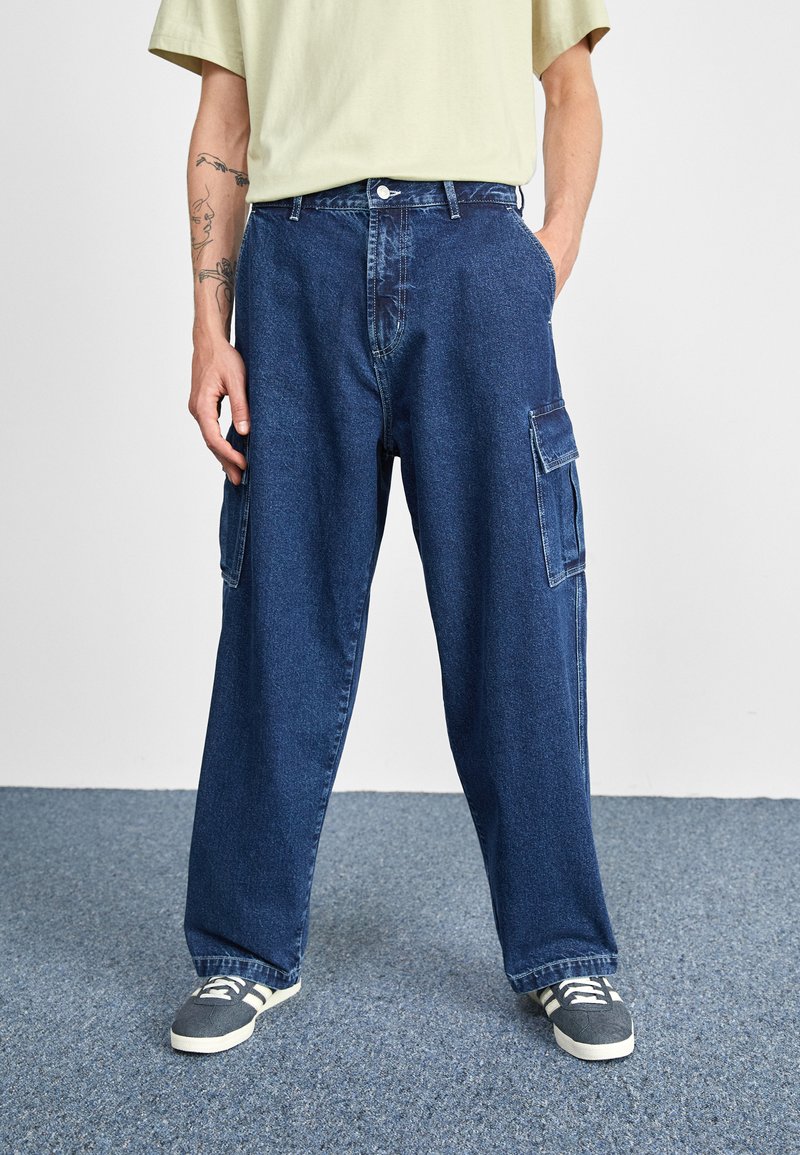 Blå denim cargo bukser med løs pasform, der har flere sidelommer og knaplukning. Sammen med grå sneakers.