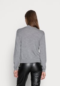 Femme aux longs cheveux bruns portant un pull gris à manches longues et un pantalon en cuir noir, vue de dos sur un fond clair uni.