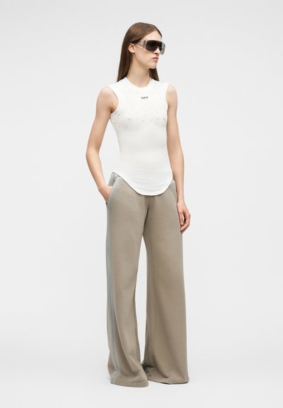 Γυναίκα με λευκό τοπ χωρίς μανίκια με το κείμενο «OFF», φαρδιά μπεζ παντελόνια και μεγάλα μαύρα γυαλιά ηλίου, στέκεται με το ένα χέρι στην τσέπη.