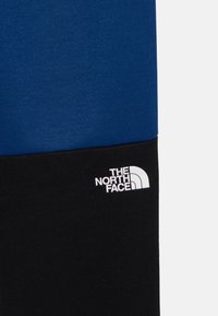 Modra in črna barvno blokirana tekstilna površina z gladko teksturo. V črnem delu se nahaja bel logotip "The North Face."