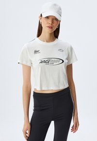 Camiseta corta blanca de algodón con estampados gráficos negros y texto. Combinada con leggings negros que tienen franjas blancas a los lados.