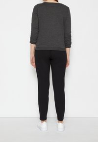 Top grigio a maniche lunghe con maniche arricciate, abbinato a pantaloni neri aderenti e sneakers bianche, visto da dietro su uno sfondo bianco.