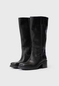 Botas de cuero negras hasta la rodilla con una textura suave, tacón ancho, costuras decorativas en blanco y un diseño ajustado.