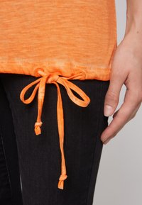 Oranges T-Shirt aus strukturiertem Stoff mit einem gerafften Saum und zwei verstellbaren Bindebändern. Über dunklen Jeans getragen, gehalten von einer Hand.