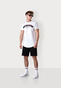 White t-shirt with black and pink print, black denim shorts, white sneakers, and patterned socks. Model poses against a neutral background.