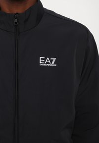 Melnā vieglā jakā ar augstu apkakli, kurai ir priekšējais rāvējslēdzis un balts izšūts EA7 Emporio Armani logo kreisajā krūtīs.