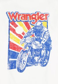 Μοτοσικλετιστής με μπλε εξοπλισμό που οδηγεί μοτοσυκλέτα μπροστά σε φόντο με ηλιαχτίδες κόκκινου, κίτρινου και μπλε χρώματος, με έντονο κόκκινο και κίτρινο κείμενο «Wrangler» πάνω από την εικόνα.