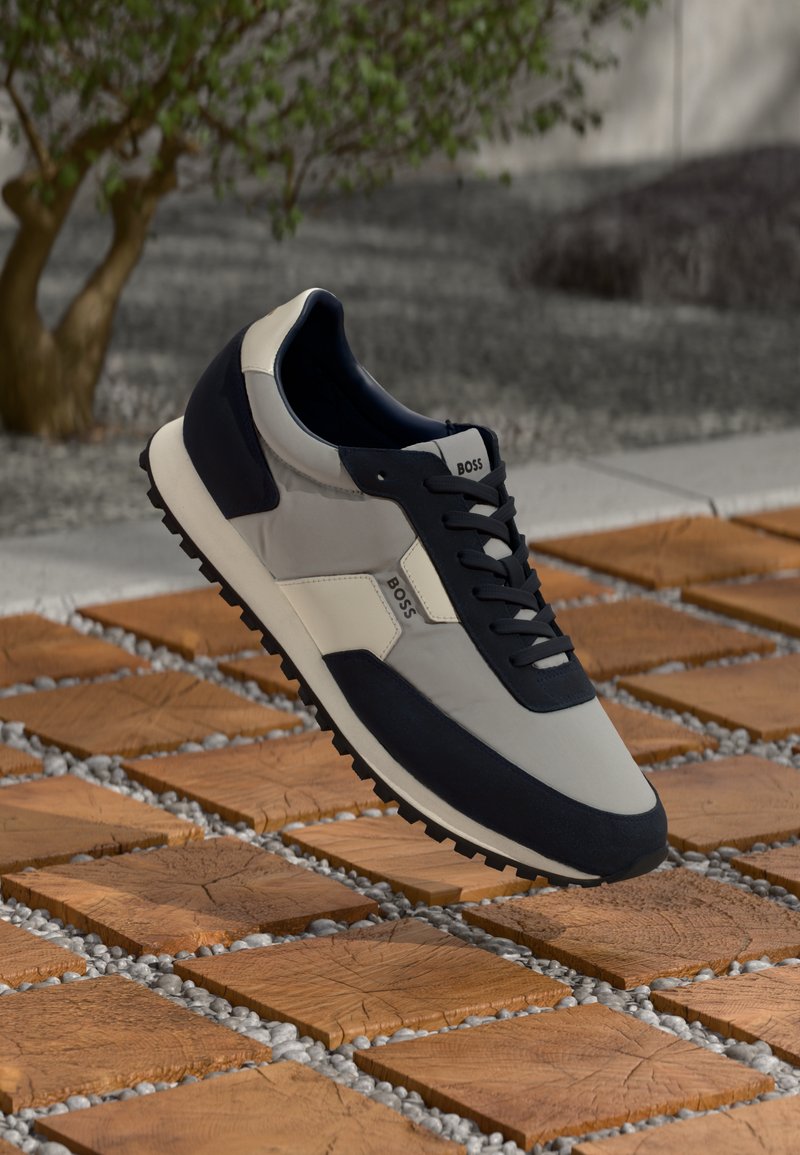 Sneakers ar pelēku, tumši zilu un baltu materiālu kombināciju. Tekstūrētā gumijas zole ar auklām un zīmola logo sānos, kas atrodas virs celiņa.