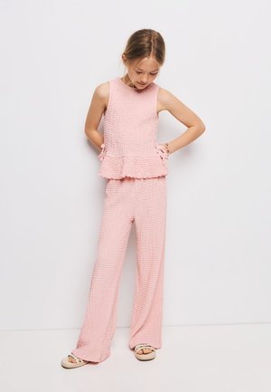 Fille portant un top sans manches rose texturé et un pantalon large assorti, debout contre un mur blanc, regardant vers le bas.