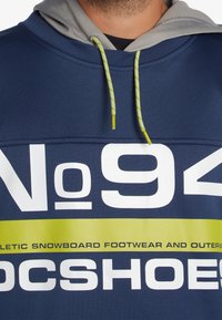 Marinblå sweatshirt med grå huva, prydd med stora vita och gula texter. Förstärkt med en texturerad, ljusgul dragsko.