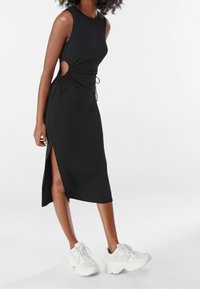 Svart ärmlös klänning med sidoutskärningar, samlad detaljer och en höga slits, i kombination med vita klumpiga sneakers. Slät textur.