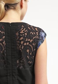 Top en dentelle noire avec un motif floral, silhouette ajustée et tissu uni contrastant au dos, doté d'une fermeture éclair.