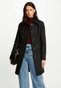 Zwarte trenchcoat met een getailleerde pasvorm, dubbelzijdig ontwerp en schouderklappen. Gecombineerd met hooggetailleerde blauwe denim jeans en een zwarte tas.