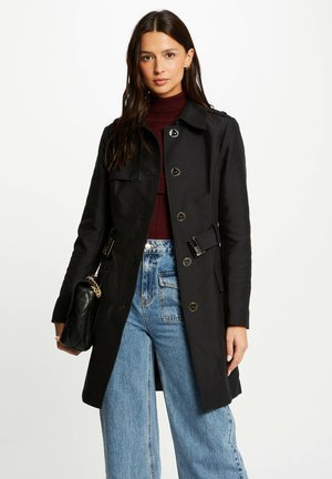 Abrigo negro con corte entallado, diseño de doble botón y hombreras. Combinado con unos vaqueros de mezclilla azul de cintura alta y un bolso negro.