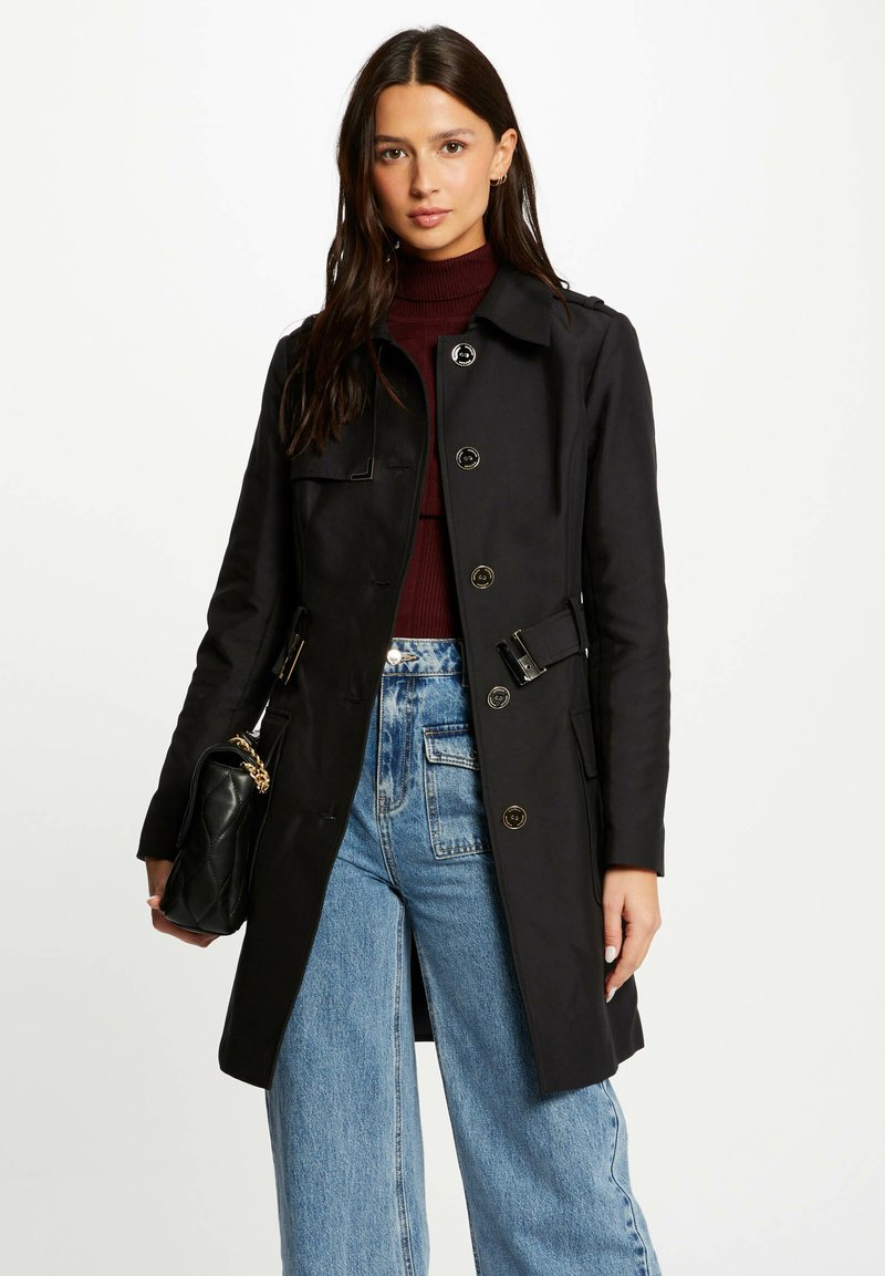 Zwarte trenchcoat met een getailleerde pasvorm, dubbelzijdig ontwerp en schouderklappen. Gecombineerd met hooggetailleerde blauwe denim jeans en een zwarte tas.