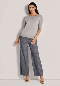 Hellgrauer Strickpullover mit kurzen Ärmeln und einem dezenten, verzierten Muster am Ausschnitt, kombiniert mit dunkel gestreiften, weiten Hosen.