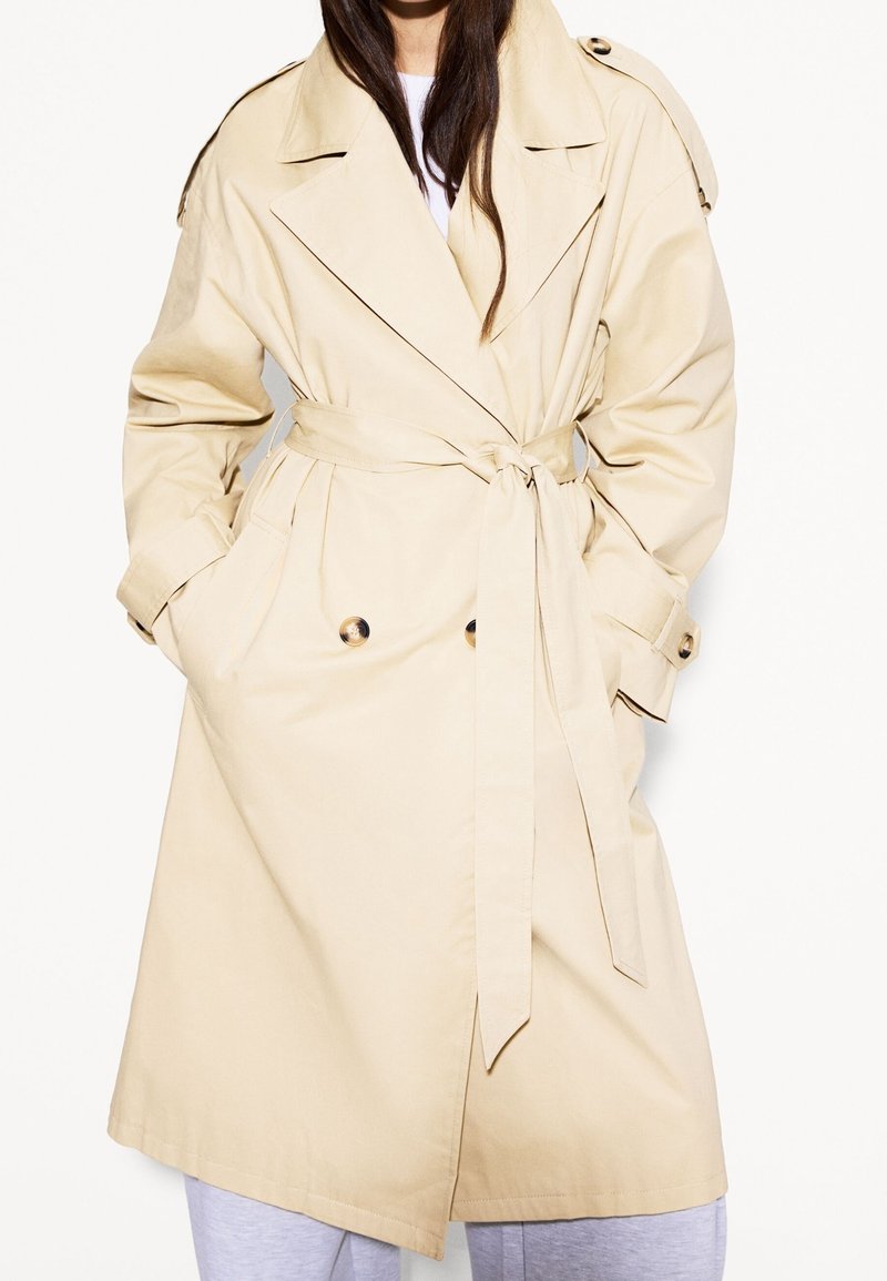Beżowy trencz z podwójnym zapięciem, szerokimi klapami, paskiem w pasie oraz kieszeniami bocznymi, wykonany z lekkiego materiału.