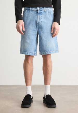Shorts en denim bleu clair, avec une coupe décontractée, des poches avant et un ourlet roulé. Associés à des chaussures noires et des chaussettes blanches.