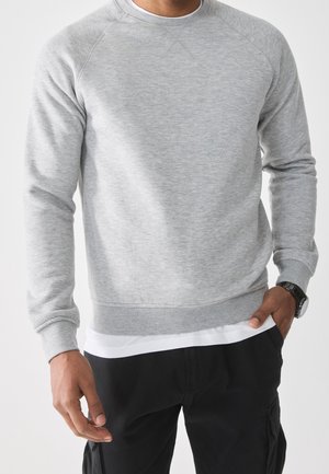 Mann trägt ein hellgraues Sweatshirt mit langen Ärmeln über einem weißen Hemd, schwarze Hose und eine schwarze Armbanduhr, steht mit einer Hand in der Tasche.
