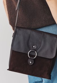 Geantă din piele neagră și piele întoarsă, cu închidere cu clapetă, detaliu cu inel de metal argintiu și textură contrastantă. Curea reglabilă.