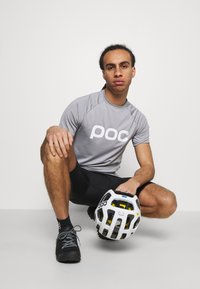 Grå atletisk t-shirt med "POC" logo, sorte shorts og sorte sko. Holder en hvid og gul cykelhjelm med ventilationshuller. Kort, tekstureret hår.