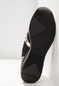 Czarna gumowa podeszwa trampka z teksturowanym bieżnikiem, wyposażona w siateczkową tkaninę i metaliczne złote akcenty na górnej części. Oznaczona napisem „MICHAEL KORS”.