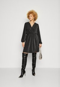 Μαύρο, λαμπερό φόρεμα με φουστάνι και μακριά μανίκια, με τσακωμένο μέσο. Συνδυάζεται με ψηλές μαύρες μπότες και κρατώντας μια μικρή ασημένια τσάντα.