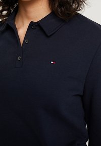 Polo blu navy in tessuto strutturato, con colletto e tre bottoni neri. Include un piccolo logo rosso, bianco e blu.