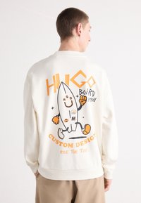 Kremfarget hettegenser med et tegneserie-surfebrettgrafikk i oransje og svart. Har teksten "CUSTOM DESIGN" og ribbestrikkede mansjetter og nederkant.