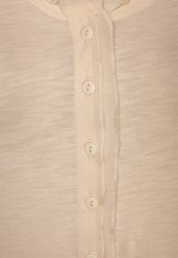 Beige topp med en texturerad tyg, som har en knappfront och sex rundade, opaka knappar. Mjuk material med en subtil lyster.
