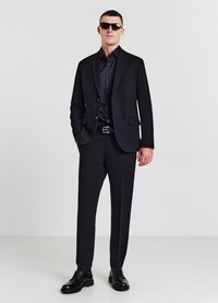 Schwarzer Anzug mit einreihigem Sakko, passendem Hemd und schmal geschnittenen Hosen. Zubehör umfasst einen schwarzen Gürtel und eine Sonnenbrille. Schwarze Lederschuhe.