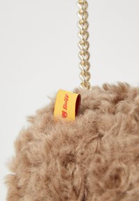 Rjav puhast torbica z zlatim verižnim pasom, ki ima rumeno oznako z napisom "Steiff." Tekstura mehke površine s poudarjenimi vlakni.