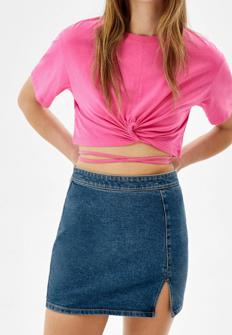 Femme portant un crop top rose noué avec des liens croisés et une mini-jupe en denim bleue avec une petite fente sur le côté.