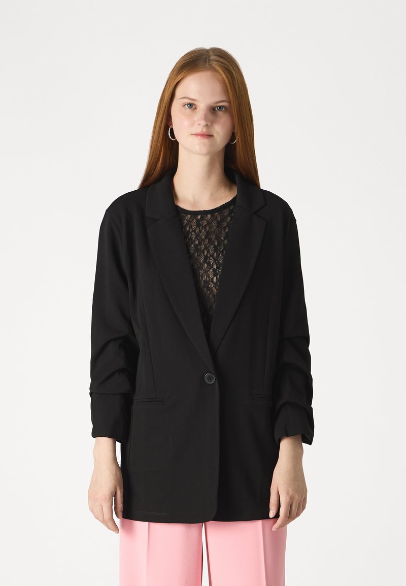 Črni blazer s strukturnim dizajnom, enojnim gumbom in zvitimi rokavi, v kombinaciji z čipkastim vrhom in rožnatimi hlačami.