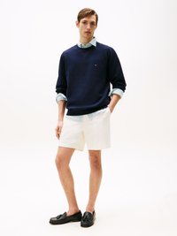 Marineblå hettegenser med rund hals og logo, lys blå skjorte med krage under, hvite shorts og svarte loafers med frynser.