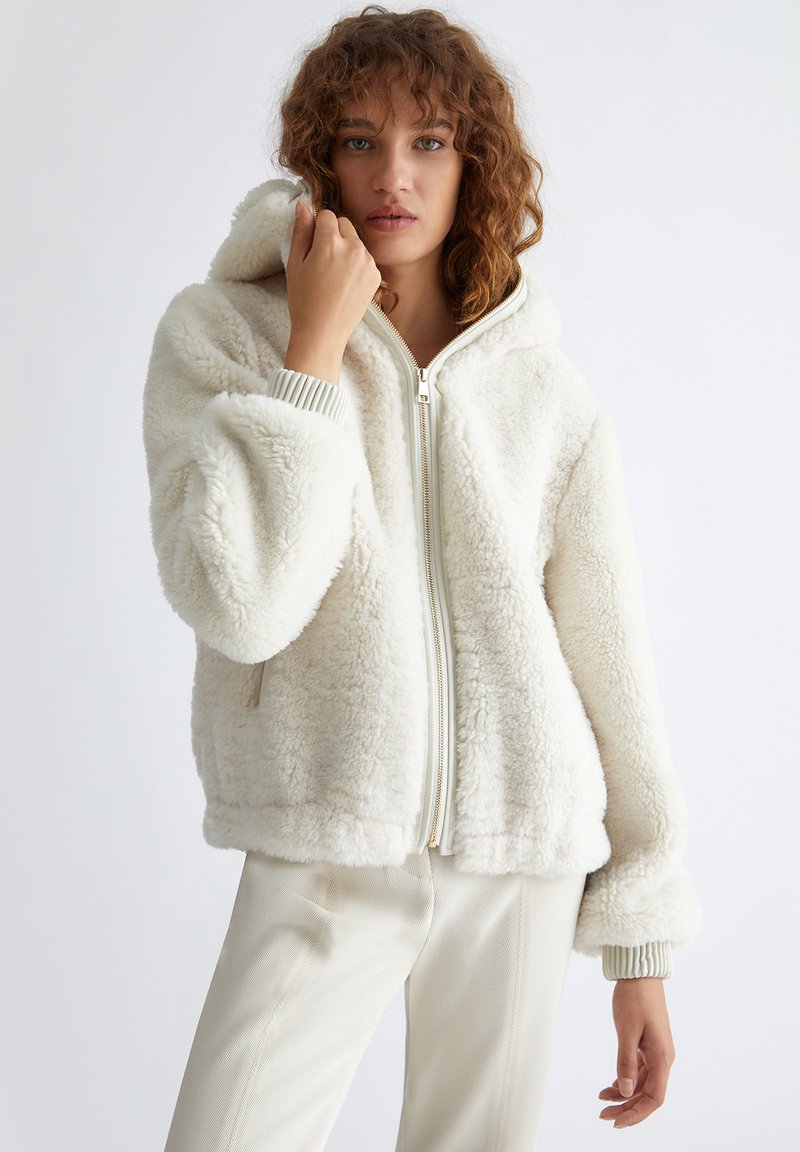 Haut à capuche zippé blanc et duveteux avec une capuche doublée, des poignets côtelés et une texture douce, présenté sur un modèle se tenant contre un fond neutre.