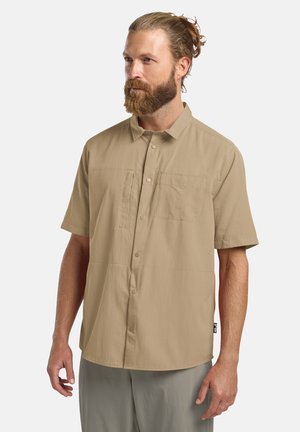 Baardige man met naar achter gebonden haar, gekleed in een beige overhemd met korte mouwen en lichtgrijze broek, staand tegen een effen achtergrond.