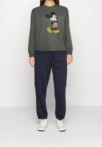 Mörkgrön sweatshirt med tryckt Mickey Mouse-design, kombinerad med marinblå sweatpants och vita sneakers. Avslappnad, bekväm stil.
