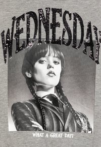T-shirt grigio con una grafica in bianco e nero di una ragazza con capelli treccia, decorata con paillettes nere che formano la scritta "MERCOLEDÌ" sopra e "CHE GRAN BEL GIORNO!" sotto.