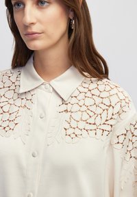 Crème blouse met een kraag, knoopsluiting en ingewikkelde bloemenborduursels over de schouders en mouwen.