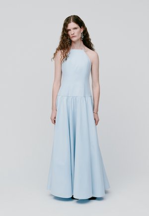 Donna con capelli lunghi e ricci che indossa un abito leggero azzurro con scollatura all'americana e lunghezza fino al pavimento, e orecchini argento, su sfondo semplice.