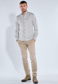 Hellgraues Button-up-Hemd mit einem dezenten Muster, beigefarbene Slim-Fit-Hose und braune Wildleder-Stiefeletten vor einem schlichten Hintergrund.