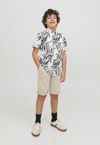 Camicia bianca a maniche corte con motivo fogliare navy, pantaloni corti beige arrotolati, calze nere e sneaker crema con suole marroni.