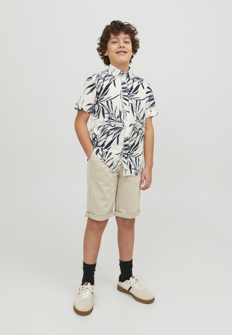 Camicia bianca a maniche corte con motivo fogliare navy, pantaloni corti beige arrotolati, calze nere e sneaker crema con suole marroni.