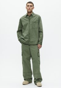 Camicia verde in cotone a maniche lunghe con chiusura a bottoni e tasca, abbinata a pantaloni cargo larghi coordinati e scarpe slip-on di colore chiaro.