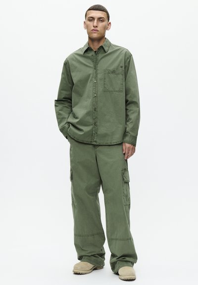 Camicia verde in cotone a maniche lunghe con chiusura a bottoni e tasca, abbinata a pantaloni cargo larghi coordinati e scarpe slip-on di colore chiaro.