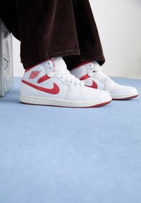Sneakers alte in pelle bianca con dettagli rossi, dotate di un logo circolare e suola texture, abbinate a pantaloni in corduroy marroni.