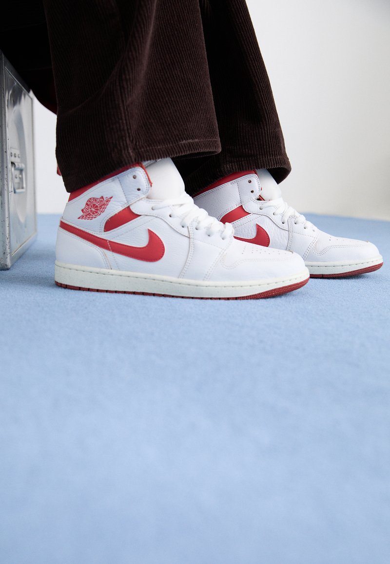 Zapatillas altas de cuero blanco con acentos rojos, que presentan un logo circular y suela texturizada, combinadas con pantalones de pana marrones.